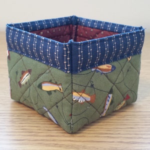 May include: A square fabric basket with a quilted design. The exterior features a green fabric with a fish pattern, while the top edge is navy blue with a dotted pattern. The interior lining is a burgundy color.