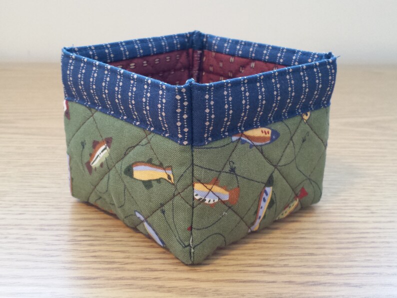 May include: A square fabric basket with a quilted design. The exterior features a green fabric with a fish pattern, while the top edge is navy blue with a dotted pattern. The interior lining is a burgundy color.