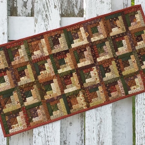 May include: A brown, green, and cream patchwork table runner with a red border. The runner is made of fabric scraps sewn together in a log cabin pattern.