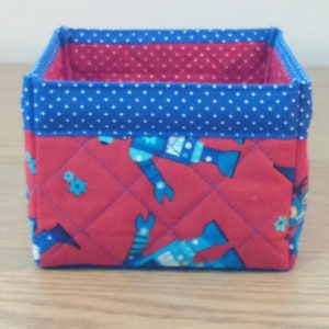 May include: A red and blue fabric storage box with a quilted design featuring robots. The box has a blue polka dot trim.