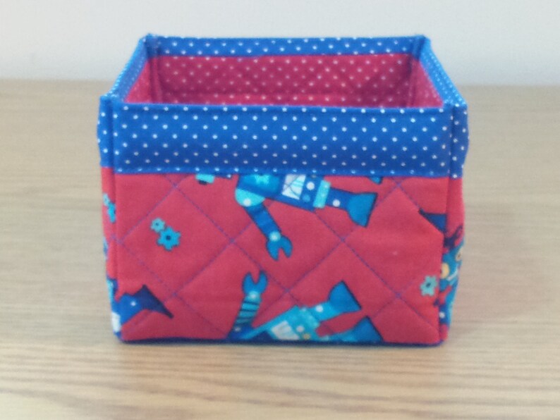 May include: A red and blue fabric storage box with a quilted design featuring robots. The box has a blue polka dot trim.