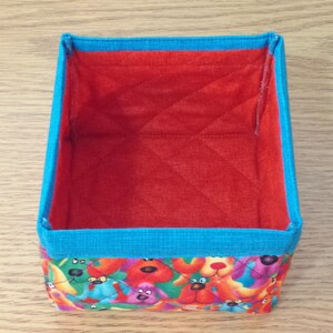 K&ouml;nnte beinhalten: Eine kleine, rechteckige Aufbewahrungsbox mit roter, gesteppter Innenauskleidung und blauem Besatz. Die Au&szlig;enseite ist mit einem bunten Stoff bezogen, der ein Muster aus Comic-Hunden zeigt.