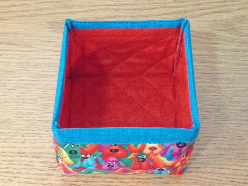 K&ouml;nnte beinhalten: Eine kleine, rechteckige Aufbewahrungsbox mit roter, gesteppter Innenauskleidung und blauem Besatz. Die Au&szlig;enseite ist mit einem bunten Stoff bezogen, der ein Muster aus Comic-Hunden zeigt.