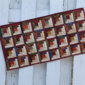 May include: A patchwork quilt runner with a log cabin pattern in shades of brown, red, cream, and green. The runner is made of various fabrics with different patterns and textures.