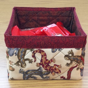 May include: A red and white quilted fabric storage box with a burgundy trim. The box is filled with red snack bars. The outside of the box features a pattern of brown and white lizards.