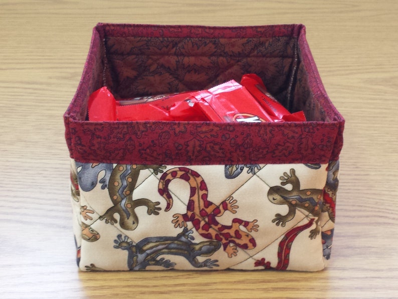 May include: A red and white quilted fabric storage box with a burgundy trim. The box is filled with red snack bars. The outside of the box features a pattern of brown and white lizards.