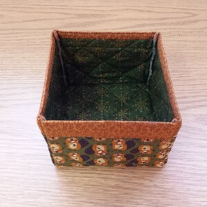 May include: A small, square, fabric storage box with a brown and orange trim. The inside of the box is lined with a green fabric with a white polka dot pattern. The outside of the box is covered in a green fabric with a brown bear pattern.