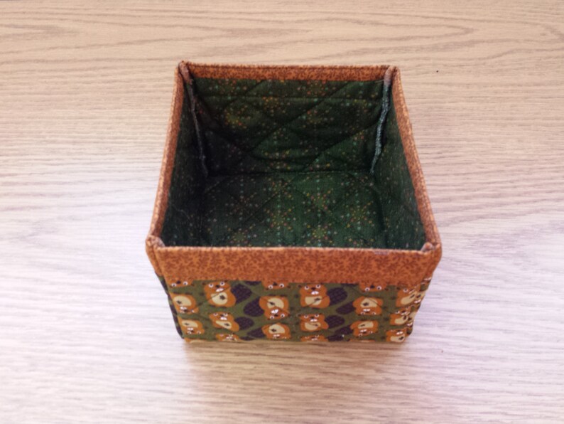 May include: A small, square, fabric storage box with a brown and orange trim. The inside of the box is lined with a green fabric with a white polka dot pattern. The outside of the box is covered in a green fabric with a brown bear pattern.
