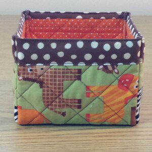 May include: A brown and green fabric storage bin with a white polka dot pattern on the top. The bin has a quilted design and features animal illustrations.