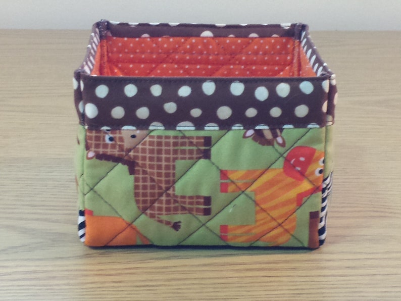 May include: A brown and green fabric storage bin with a white polka dot pattern on the top. The bin has a quilted design and features animal illustrations.