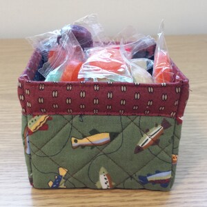May include: A small, square fabric basket filled with wrapped candies. The basket has a quilted green exterior with a fish pattern and a burgundy top edge. The candies are various colors and shapes.