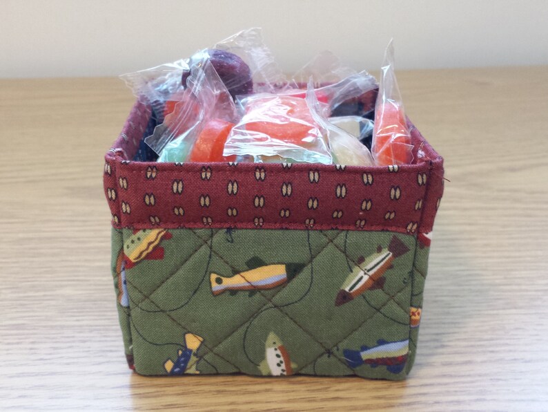 May include: A small, square fabric basket filled with wrapped candies. The basket has a quilted green exterior with a fish pattern and a burgundy top edge. The candies are various colors and shapes.