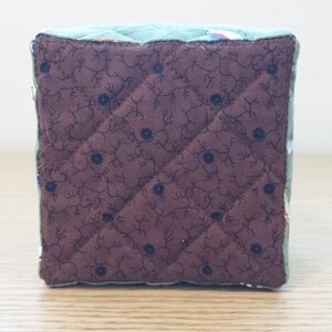 May include: A quilted cube with a brown floral patterned bottom and a teal top. The brown fabric features a diamond pattern with small black flower accents. The edges are trimmed with a teal fabric.