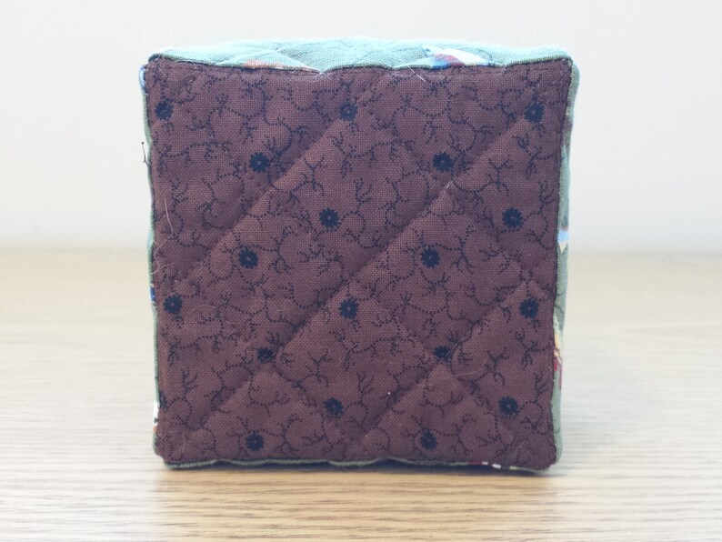 May include: A quilted cube with a brown floral patterned bottom and a teal top. The brown fabric features a diamond pattern with small black flower accents. The edges are trimmed with a teal fabric.