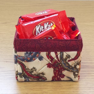 May include: A red fabric storage basket with a quilted design featuring a pattern of lizards. The basket is filled with several red and white Kit Kat candy bar wrappers.