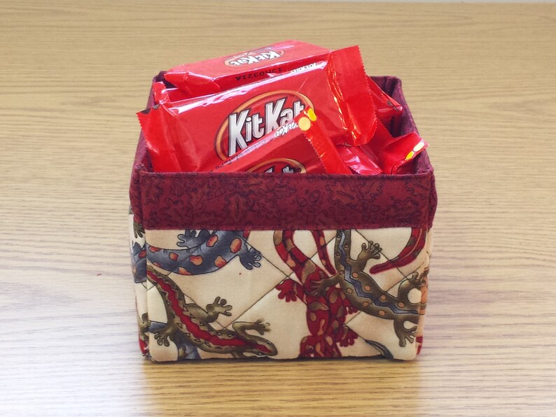 May include: A red fabric storage basket with a quilted design featuring a pattern of lizards. The basket is filled with several red and white Kit Kat candy bar wrappers.