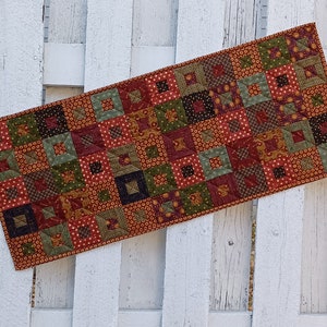 May include: A patchwork table runner with a variety of red, green, brown, and gold fabrics. The runner is made of squares and rectangles, and the fabric patterns include polka dots, floral prints, and geometric designs.