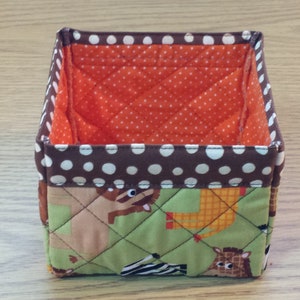 May include: A square-shaped fabric storage bin with a brown and white polka dot trim. The bin is lined with orange fabric with white polka dots. The exterior of the bin features a green quilted fabric with a pattern of animals, including a zebra, giraffe, and lion.