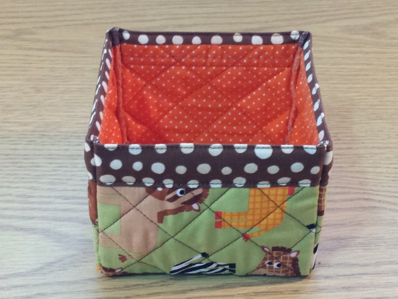 May include: A square-shaped fabric storage bin with a brown and white polka dot trim. The bin is lined with orange fabric with white polka dots. The exterior of the bin features a green quilted fabric with a pattern of animals, including a zebra, giraffe, and lion.