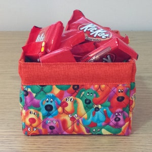 K&ouml;nnte beinhalten: Ein roter Stoffaufbewahrungskorb mit einem gesteppten Design mit bunten Comic-Hunden. Der Korb ist gef&uuml;llt mit roten und wei&szlig;en S&uuml;&szlig;igkeitenverpackungen mit dem Text "Kit Kat Crisp Wafers".
