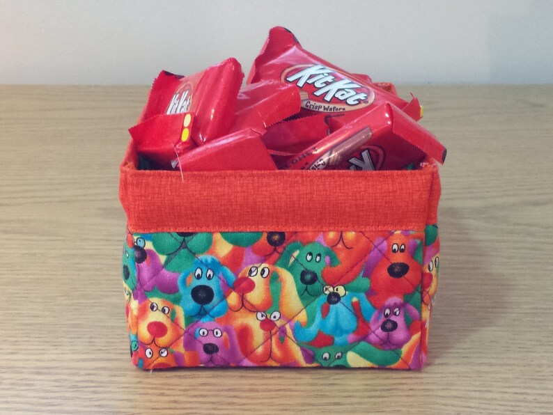 K&ouml;nnte beinhalten: Ein roter Stoffaufbewahrungskorb mit einem gesteppten Design mit bunten Comic-Hunden. Der Korb ist gef&uuml;llt mit roten und wei&szlig;en S&uuml;&szlig;igkeitenverpackungen mit dem Text "Kit Kat Crisp Wafers".