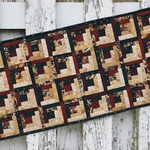 May include: A rectangular table runner with a patchwork design featuring squares of beige, brown, and green fabric. The fabric has a variety of patterns, including floral, geometric, and plaid.