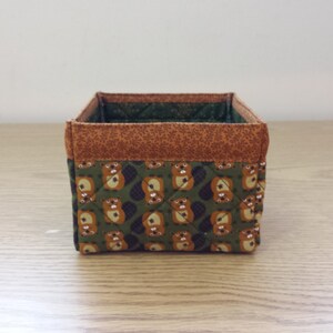 May include: A small, square storage basket made of fabric with a brown and green pattern of animals. The basket has a brown top and a green bottom.
