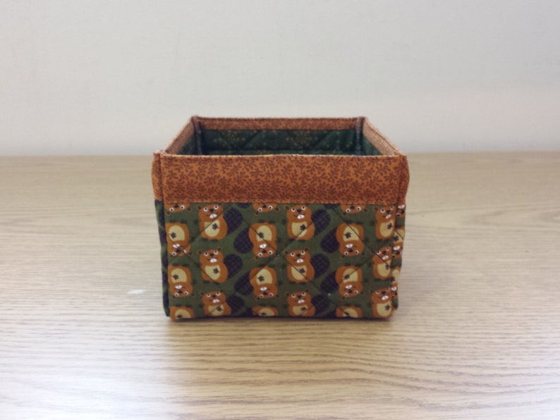 May include: A small, square storage basket made of fabric with a brown and green pattern of animals. The basket has a brown top and a green bottom.