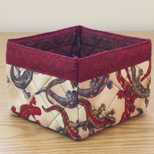 May include: A square-shaped fabric storage basket with a red trim. The basket is decorated with a pattern of colorful lizards on a cream background.