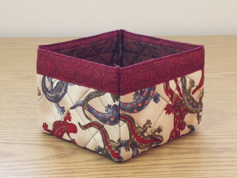 May include: A square-shaped fabric storage basket with a red trim. The basket is decorated with a pattern of colorful lizards on a cream background.