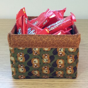 May include: A brown and green fabric storage basket with a quilted pattern of beavers. The basket is filled with red and white candy bars.