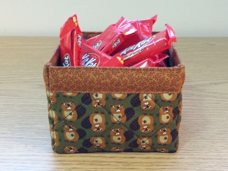 May include: A brown and green fabric storage basket with a quilted pattern of beavers. The basket is filled with red and white candy bars.