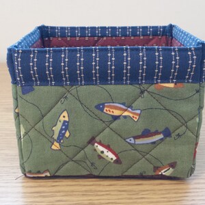 May include: A small, square fabric storage basket. The exterior features a green fabric with a fish pattern and quilted stitching. The top edge is trimmed with a blue fabric with a white dotted pattern. The interior is lined with a burgundy fabric.