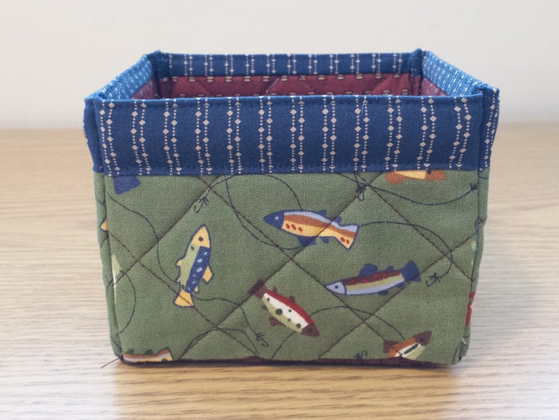 May include: A small, square fabric storage basket. The exterior features a green fabric with a fish pattern and quilted stitching. The top edge is trimmed with a blue fabric with a white dotted pattern. The interior is lined with a burgundy fabric.