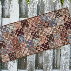 May include: A brown, blue, and cream patchwork quilt table runner with a geometric pattern. The runner is hanging on a white picket fence.