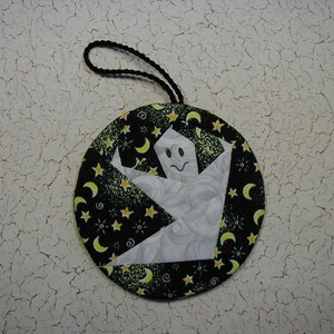 May include: A round, black fabric ornament with a white ghost and yellow stars and crescent moons. The ornament has a black string loop for hanging.