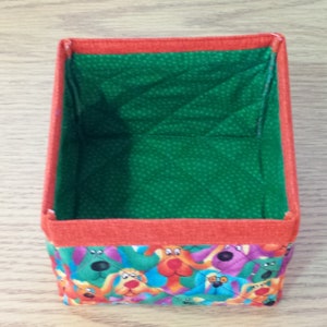 K&ouml;nnte beinhalten: Eine kleine, quadratische Aufbewahrungsbox mit gr&uuml;ner Stoffauskleidung und einem bunten Stoff&auml;u&szlig;eren mit einem Muster aus Comic-Hunden. Die Box hat einen roten Rand um die obere Kante.