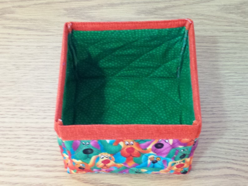 K&ouml;nnte beinhalten: Eine kleine, quadratische Aufbewahrungsbox mit gr&uuml;ner Stoffauskleidung und einem bunten Stoff&auml;u&szlig;eren mit einem Muster aus Comic-Hunden. Die Box hat einen roten Rand um die obere Kante.