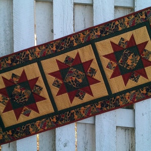 May include: A table runner with a green, gold, and red starburst pattern. The runner has a border of green leaves and a red outer edge.