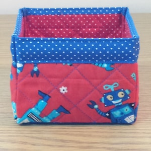 May include: A red and blue fabric storage box with a quilted design featuring robots. The box has a blue polka dot trim.