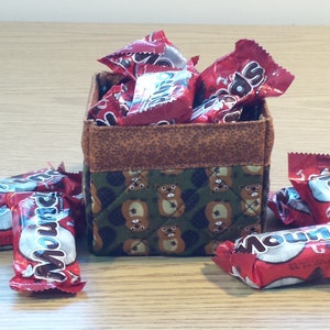 May include: A brown and green fabric storage box with a quilted pattern of bears. The box is filled with red and white candy wrappers. The candy wrappers have the word "Mounds" printed on them.