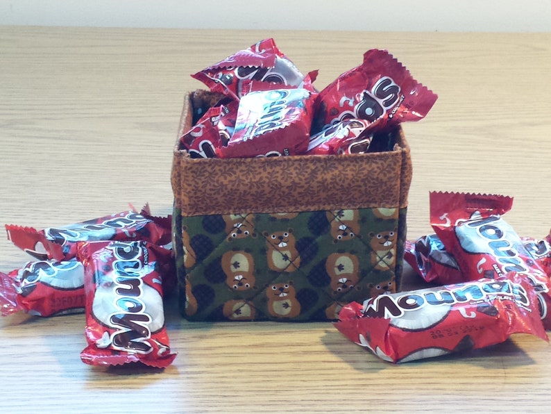 May include: A brown and green fabric storage box with a quilted pattern of bears. The box is filled with red and white candy wrappers. The candy wrappers have the word "Mounds" printed on them.