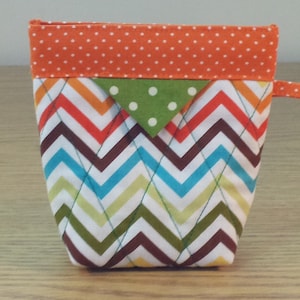 May include: A small, quilted zippered pouch with a chevron pattern in shades of brown, green, blue, orange, and yellow. The pouch has a top flap with orange polka dots and a green triangle with white polka dots.