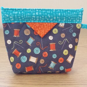 May include: A small gray zippered pouch with a turquoise top and orange accent. The pouch is decorated with a repeating pattern of buttons, spools of thread, and needles.