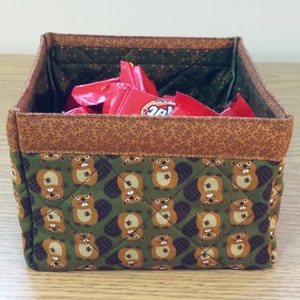 May include: A green fabric storage bin with a brown trim and a pattern of beavers. The bin is filled with red and white candy wrappers.