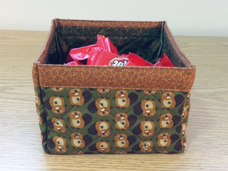 May include: A green fabric storage bin with a brown trim and a pattern of beavers. The bin is filled with red and white candy wrappers.