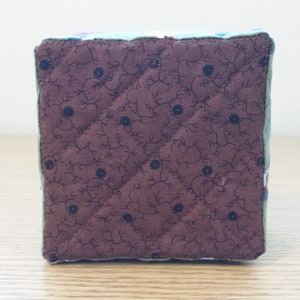May include: A square, quilted fabric cube with a brown floral pattern on one side. The cube has a light green border and is likely handmade. The brown fabric features a repeating pattern of small blue flowers.