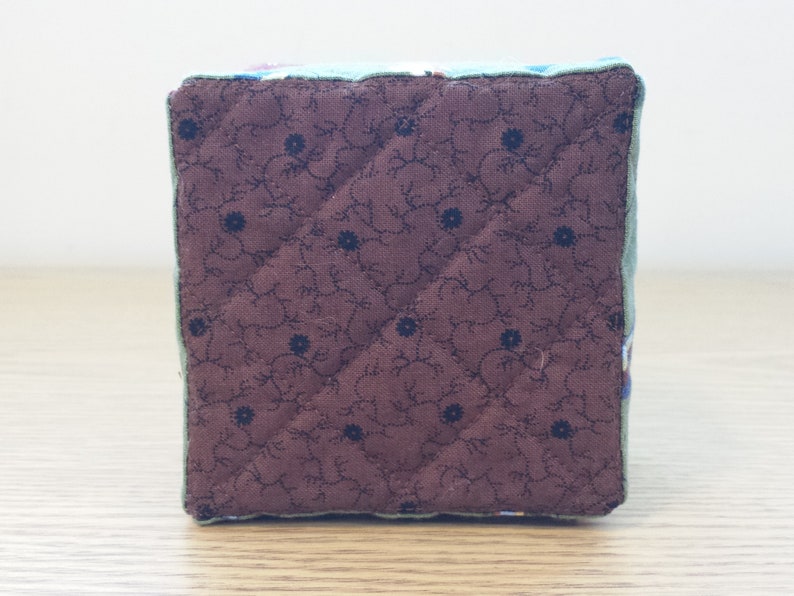 May include: A square, quilted fabric cube with a brown floral pattern on one side. The cube has a light green border and is likely handmade. The brown fabric features a repeating pattern of small blue flowers.