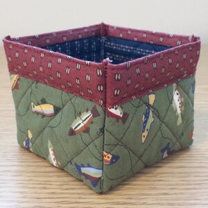 May include: A square, quilted fabric basket with a fish pattern. The basket features a green fabric with colorful fish, a burgundy top band with a small white pattern, and a blue interior. The basket is likely handmade.