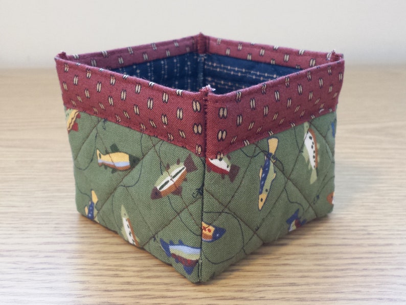 May include: A square, quilted fabric basket with a fish pattern. The basket features a green fabric with colorful fish, a burgundy top band with a small white pattern, and a blue interior. The basket is likely handmade.